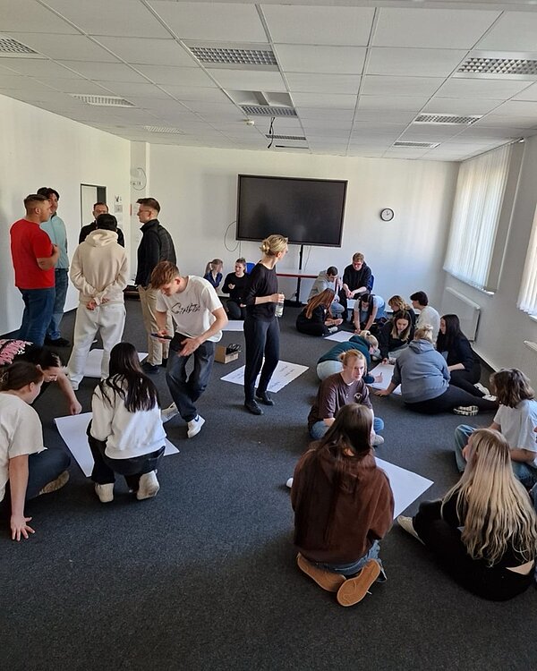 Theaterbesuch und Workshop Die Auszubildenden stehen und sitzen während des Theaterworkshops gemeinsam im Raum und arbeiten in Gruppen.