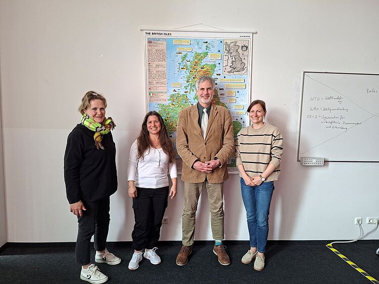Vier lächelnde Erwachsene posieren vor einer großen Wandkarte der Britischen Inseln. Von links nach rechts: Eine Frau mit einem schwarzen Pullover und bunten Schal, eine Frau mit weißem Oberteil und schwarzen Hosen, ein Mann in einem braunen Sakko und hellen Hosen, und eine Frau mit einem gestreiften Pullover und Jeans. Rechts im Bild steht ein Whiteboard mit handschriftlichen Notizen.