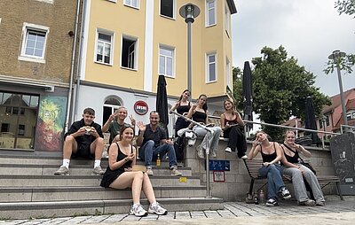 Eine Gruppe von neun Personen sitzt entspannt auf den Stufen vor einem gelben Gebäude mit weißem Fensterrahmen. Im Hintergrund ist ein Sushi-Restaurant zu erkennen. Einige Personen zeigen Peace-Zeichen oder lächeln in die Kamera. Die Szene wirkt locker und gesellig, mit urbanem Flair durch die umliegenden Gebäude und Straßenlaternen.
