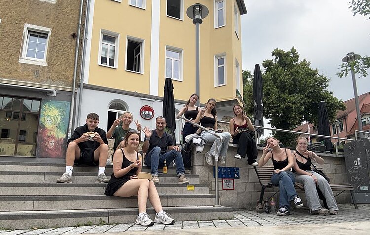 Eine Gruppe von neun Personen sitzt entspannt auf den Stufen vor einem gelben Gebäude mit weißem Fensterrahmen. Im Hintergrund ist ein Sushi-Restaurant zu erkennen. Einige Personen zeigen Peace-Zeichen oder lächeln in die Kamera. Die Szene wirkt locker und gesellig, mit urbanem Flair durch die umliegenden Gebäude und Straßenlaternen.