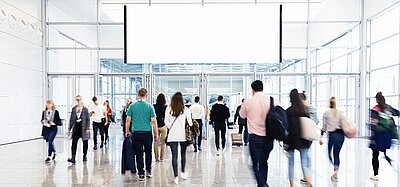 Im lichtdurchfluteten Flur oder der Eingangshalle eines Messegebäudes laufen verschiedene Menschen hektisch hin und her. Durch die Langzeitbelichtung kann man die Bewegung gut erkennen.