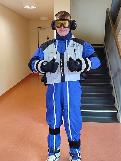 Auszubildender trägt Kopfhörer, Skibrille, Schneehandschuhe, Ganzkörperanzug mit beschwerender Weste, Halskrause und Gewichten an den Beinen. 