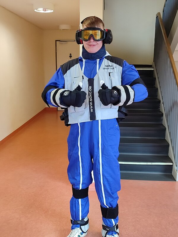 „Demenzparcours“ Auszubildender trägt Kopfhörer, Skibrille, Schneehandschuhe, Ganzkörperanzug mit beschwerender Weste, Halskrause und Gewichten an den Beinen.