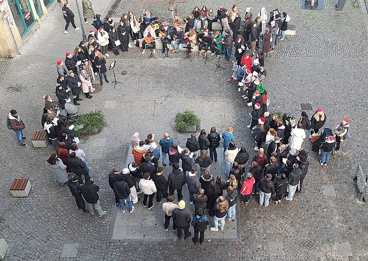 Luftaufnahme eines weihnachtlichen Singens: Eine große Gruppe von Menschen steht in einem weiten Kreis auf dem gepflasterten Marktplatz, in der Mitte die Gitarrengruppe, rundherum Zuhörer*innen mit Mützen und Winterjacken.