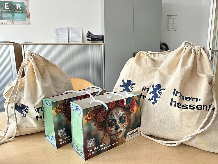 Mehrere Stoffbeutel mit „inhessen“-Aufdruck und bunte Werbegeschenktüten stehen auf einem Tisch in einem Innenraum.