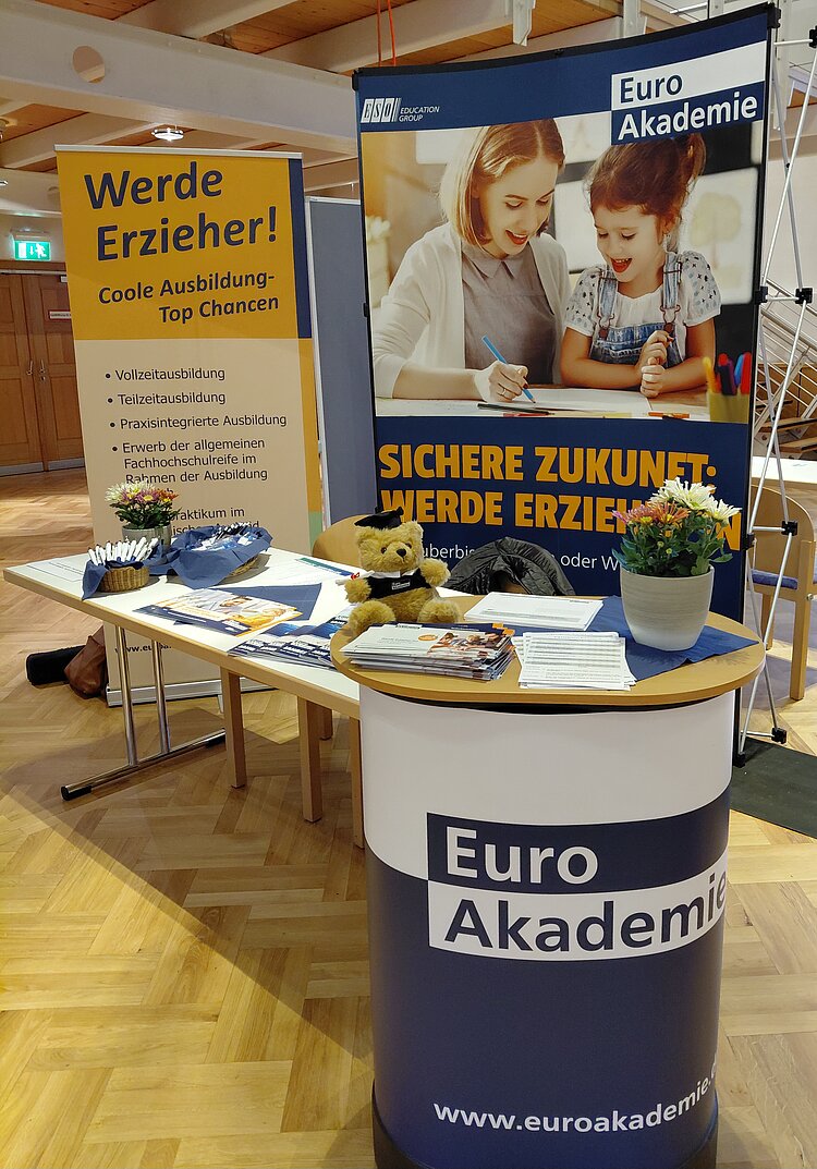 Messestand der Euro Akademie Tauberbischofsheim, geschmückt mit Blumentöpfen und einem Teddybär.