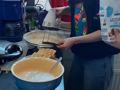 Eine Person steht in einer Küche und bereitet Waffelteig zu. Sie hält eine Packung Mehl in der Hand und lächelt in die Kamera. Auf dem Küchentisch stehen eine Schüssel mit Teig, eine Waffelmaschine und ein Teller mit fertigen Waffeln.