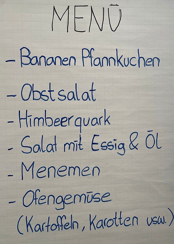 Handschriftlich gestaltete Menütafel auf weißem Papier mit blauer Schrift. Überschrift: 'MENÜ'. Darunter eine Liste mit sechs Gerichten, jeweils mit einem Bindestrich eingeleitet: Bananen Pfannkuchen, Obstsalat, Himbeerquark, Salat mit Essig & Öl, Menemen, Ofengemüse. In Klammern unter Ofengemüse steht: Kartoffeln, Karotten usw.
