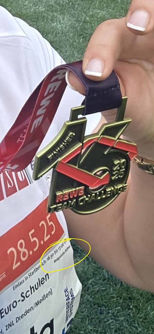 Nahaufnahme einer Hand, die die REWE Team Challenge 2025 Finisher-Medaille hält. Im Hintergrund sieht man das Startnummernfeld mit Veranstaltungsinfos.