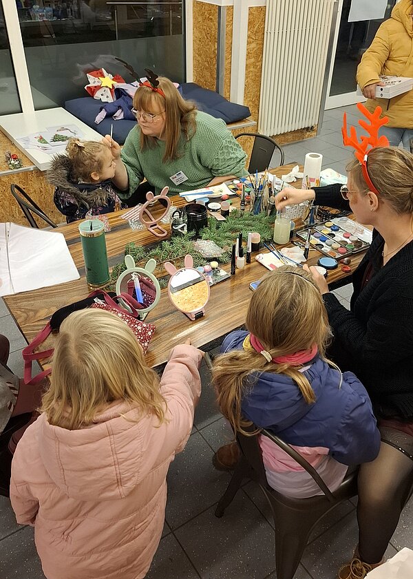 Mehrere Kinder sitzen an einem Basteltisch, während zwei Betreuerinnen mit Rentier-Haarreifen Gesichter bemalen; der Tisch ist voll mit Schminkfarben, Pinseln, Spiegeln und weihnachtlicher Dekoration.