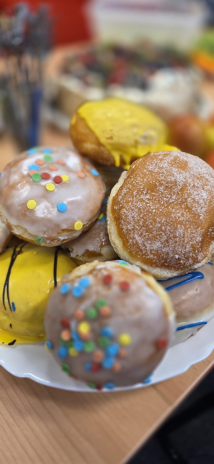 Nahaufnahme eines Tellers mit mehreren Berlinern/Krapfen: einige mit gelbem Zuckerguss und Schokostreifen, andere mit weißer Glasur und bunten Streuseln sowie ein gezuckerter Berliner.