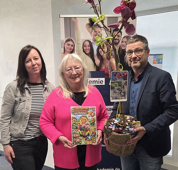 Auf dem Bild stehen drei Personen, links zwei Frauen und rechts ein Mann, mit einem Blumenstrauß in der Hand und lächeln in die Kamera.