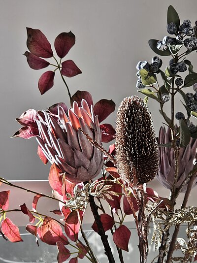 Ein kunstvolles Arrangement aus getrockneten Blumen und Pflanzen, darunter ein großer Protea, ein Banksia-Zapfen und verschiedene Blätter in warmen Rottönen und silbrigem Glanz, vor einem neutralen Hintergrund.