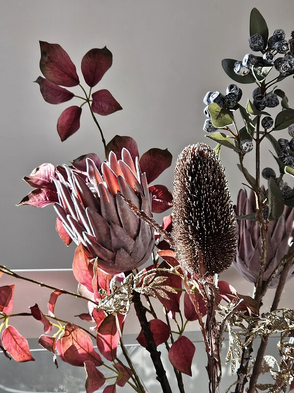 Die besten Neujahrswünsche Ein kunstvolles Arrangement aus getrockneten Blumen und Pflanzen, darunter ein großer Protea, ein Banksia-Zapfen und verschiedene Blätter in warmen Rottönen und silbrigem Glanz, vor einem neutralen Hintergrund.