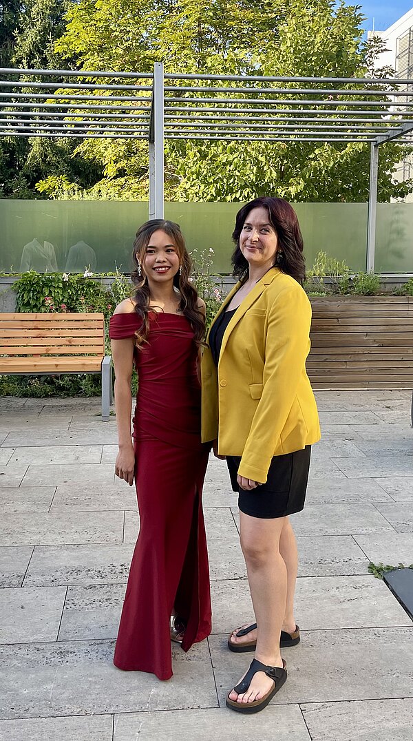 In einem Hof mit Bäumen und Bänken im Hintergrund stehen eine junge Frau in dunkelrotem Kleid und eine Dozentin in schwarzem Kleid und gelbem Blazer.