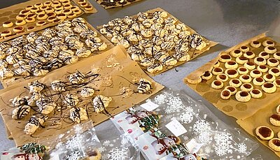 Viele Plätzchen mit Schokoguss und Husarenkrapfen mit Marmelade liegen auf mehreren Blättern Backpapier auf einem Metalltisch; daneben liegen kleine Plastiktüten mit Schneemannmotiven.