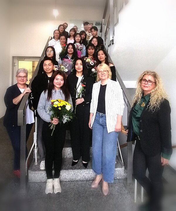 Die Gruppe von Absolventinnen und Absolventen der Pflegefachkraft-Ausbildung der Euro Akademie Bitterfeld-Wolfen steht auf einer Treppe im Schulgebäude. Viele von ihnen halten Blumensträuße und lächeln, gemeinsam mit ihren Lehrkräften, die ebenfalls für das Erinnerungsfoto posieren, in die Kamera.