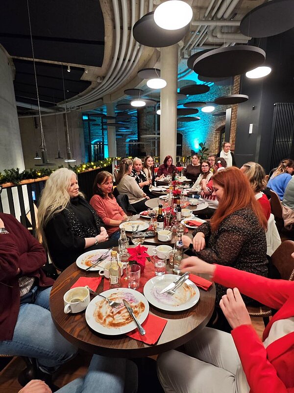 Eine Gruppe von Menschen sitzt an einem langen Tisch in einem gemütlichen Raum mit Ziegelsteinwänden und moderner Beleuchtung. Die Personen sind in Gespräche vertieft und essen gemeinsam. Auf dem Tisch stehen Teller, Gläser, Flaschen und Servietten. Die Atmosphäre wirkt gesellig und entspannt.