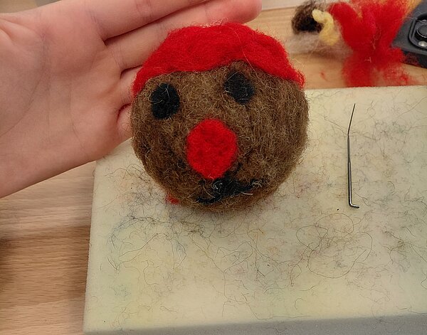 Gefilzte Kugel mit Gesicht, roter Mütze und roter Nase liegt auf einer Filzunterlage, daneben eine Hand mit ausgestreckter Innenfläche.