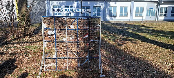 Auf einer laubbedeckten Wiese vor einem weißen Gebäude steht ein selbstgebautes Insektenhotel, gefüllt mit Laub; darüber ein Schild, auf dem steht "Euro Akademie RL".