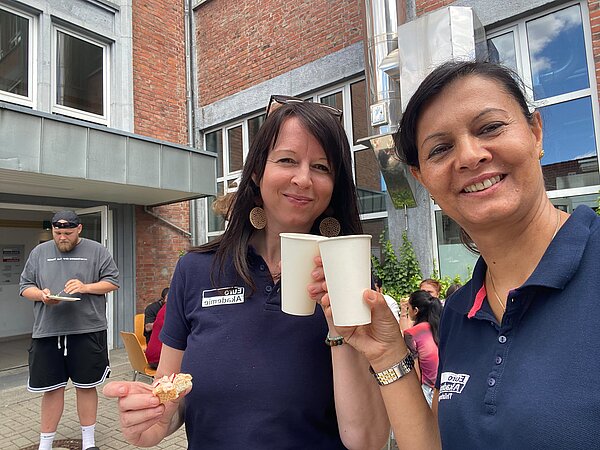 Zwei Mitarbeiterinnen der Euro Akademie stoßen bei einem Sommerfest im Innenhof mit Pappbechern an und lächeln in die Kamera.