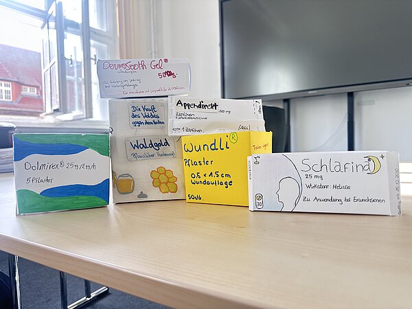 Mehrere selbst gestaltete Modellverpackungen von Arzneimitteln stehen auf einem Tisch. Zu sehen sind unter anderem Verpackungen für Pflaster, Gel, Hustensaft und ein Schlafmittel.