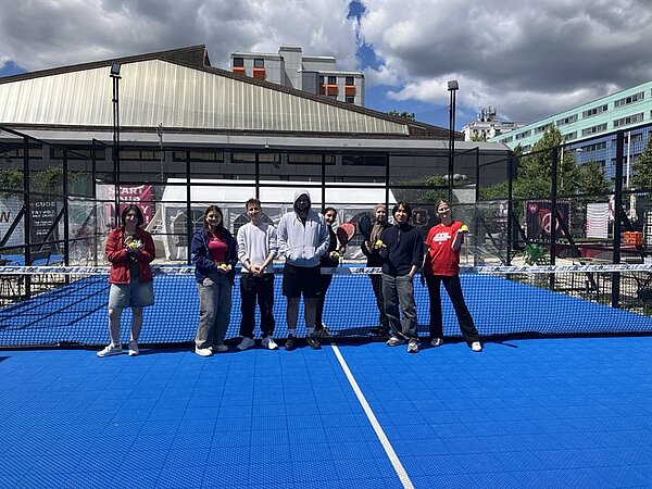 Die Gruppe der Auszubildenden steht auf einem blauen Padelplatz vor einem Netz. Im Hintergrund sind ein modernes Gebäude mit Flachdach, ein mehrstöckiges Wohnhaus und Bäume zu sehen. Die Personen tragen sportliche Freizeitkleidung, einige halten Padel-Schläger in der Hand. Der Himmel ist teils bewölkt, Sonnenlicht fällt auf den Platz.