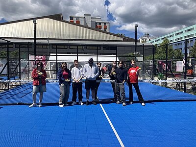 Die Gruppe der Auszubildenden steht auf einem blauen Padelplatz vor einem Netz. Im Hintergrund sind ein modernes Gebäude mit Flachdach, ein mehrstöckiges Wohnhaus und Bäume zu sehen. Die Personen tragen sportliche Freizeitkleidung, einige halten Padel-Schläger in der Hand. Der Himmel ist teils bewölkt, Sonnenlicht fällt auf den Platz.