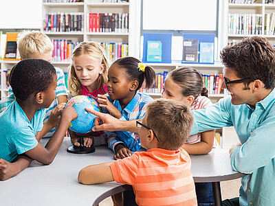 Ein Lehrer und mehrere Grundschulkinder sitzen um einen Tisch in einer Bibliothek und betrachten gemeinsam einen Globus. Die Kinder zeigen interessiert auf verschiedene Teile des Globus, während der Lehrer erklärt.