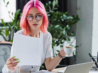 Eine Person mit pinkfarbenen Haaren und Brille sitzt an einem Schreibtisch und hält eine Tasse Kaffee in der einen Hand und ein Blatt Papier in der anderen. Im Hintergrund sind Pflanzen und ein Laptop zu sehen.