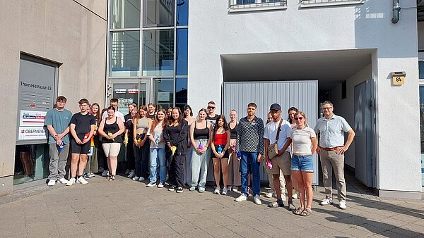 Erster Schultag: Alle an die Zuckertüten In einem Innenhof steht eine Gruppe junger Mädchen und Jungen in sommerlicher Kleidung, hinter ihnen ist ein halb geöffnetes Tor und eine geschlossene Glastür, die in ein Treppenhaus führt. Die jungen Menschen halten kleine Schultüten/Zuckertüten in den Händen. Einige ältere Personen, offenbar Lehrerinnen und Lehrer, haben keine solchen Tüten in der Hand. An der linken Hauswand sieht man eine Tafel, auf der Thomasstraße 83 steht und Logos der dort befindlichen Unternehmen sind.