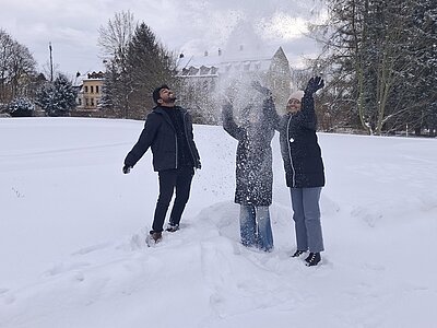 Die drei Auszubildenden stehen auf einer schneebedeckten Wiese und werfen gemeinsam Schnee in die Luft. Sie tragen dunkle Winterjacken, lange Hosen und feste Schuhe. Im Hintergrund sind Bäume und ein großes Gebäude mit spitzem Dach zu sehen. Die Szene wirkt winterlich und lebendig, passend zum Kontext ‚Es schneit, es schneit…‘.