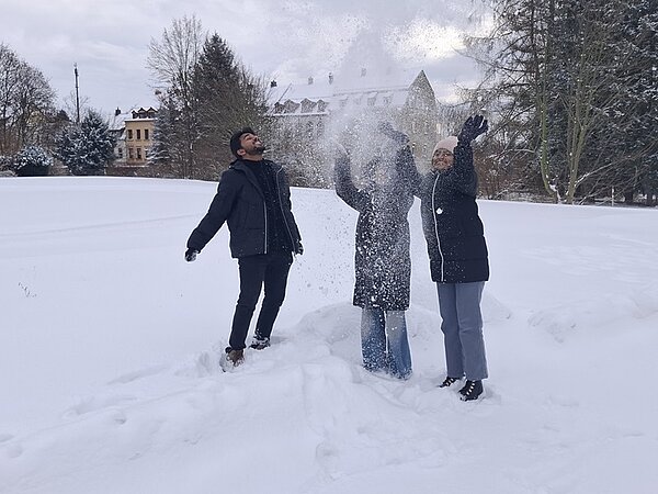 Die drei Auszubildenden stehen auf einer schneebedeckten Wiese und werfen gemeinsam Schnee in die Luft. Sie tragen dunkle Winterjacken, lange Hosen und feste Schuhe. Im Hintergrund sind Bäume und ein großes Gebäude mit spitzem Dach zu sehen. Die Szene wirkt winterlich und lebendig, passend zum Kontext ‚Es schneit, es schneit…‘.