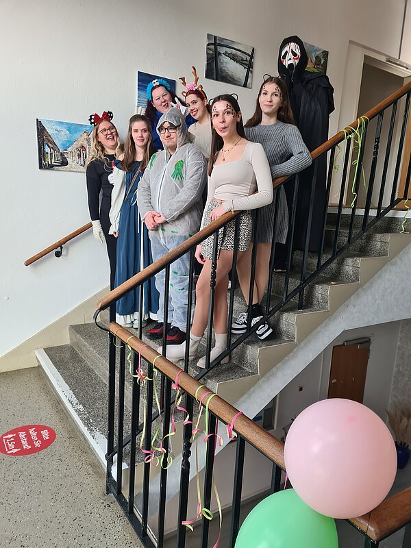 Helau und Alaaf in Rochlitz Eine Gruppe von acht Menschen in kreativen Kostümen steht auf einer Treppe in einem geschmückten Flur. Einige tragen Tier-Accessoires wie Geweih oder Katzenohren, andere sind in fantasievollen Outfits oder einem gruseligen "Scream"-Kostüm gekleidet. Die Treppe ist mit bunten Luftschlangen und Luftballons dekoriert.