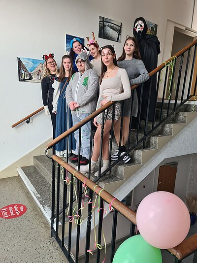 Eine Gruppe von acht Menschen in kreativen Kostümen steht auf einer Treppe in einem geschmückten Flur. Einige tragen Tier-Accessoires wie Geweih oder Katzenohren, andere sind in fantasievollen Outfits oder einem gruseligen "Scream"-Kostüm gekleidet. Die Treppe ist mit bunten Luftschlangen und Luftballons dekoriert.