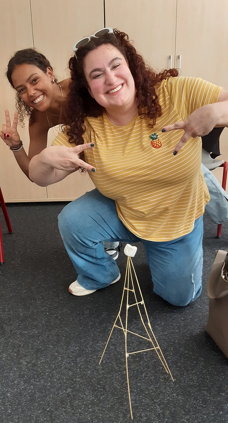 Die “Marshmallow Challenge 2.0” Lächelnd posieren zwei Schülerinnen vor ihrer gebauten Marshmallow-Konstruktion aus langen Spaghetti und einem Marshmallow auf der Spitze.