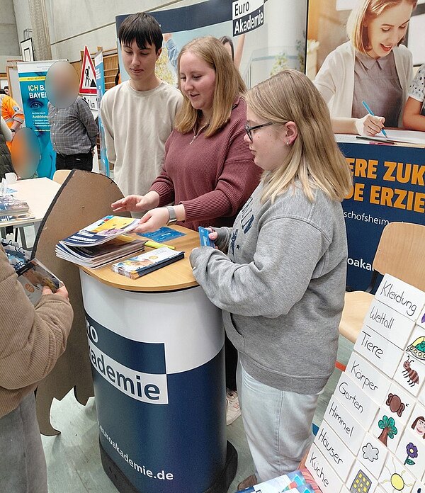 Auf dem Bild stehen drei  Auszubildende der Euro Akademie Tauberbischofsheim eng beieinander am Stehpult des Euro-Akademie-Messestandes auf der Ausbildungsmesse. Hinter ihnen befinden sich Roll-Ups mit dem Logo der Euro Akademie sowie dem Slogan ‚Werde Erzieher!‘. Vor der Gruppe steht ein Besucher, den man nicht ganz sieht, und lässt sich von den Auszubildenden beraten.