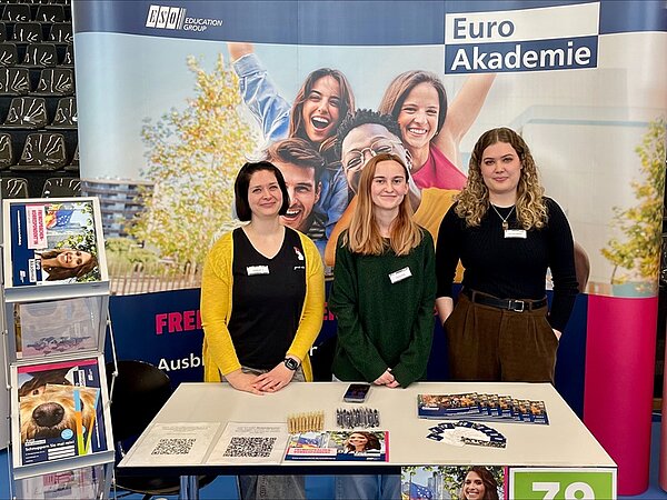 Zwei Lehrerinnen und eine Auszubildende stehen lächelnd am Messestand, hinter ihnen ein großer Euro Akademie-Aufsteller, links Informationsbroschüren und auf dem Tisch liegen Flyer und Stifte.
