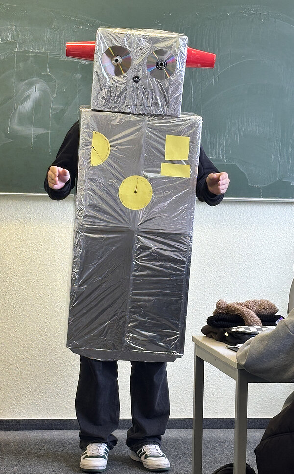 Detailaufnahme des Schülers im Roboterkostüm. Die silberne Verkleidung besteht aus Kartons mit CDs als Augen, roten Bechern als Ohren und gelben Papier-Elementen als Knöpfe.