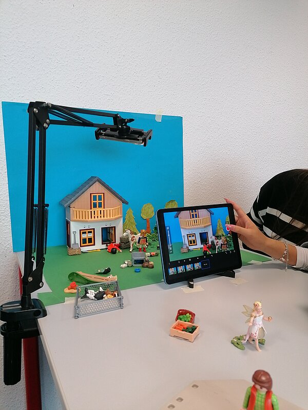 MeKoSax-Projekt „Trickfilmproduktion“ Eine Schülerin filmt ein aus Playmobil gebautes Haus mit Garten und Bäumen, welche auf einem grünen Stück Papier stehen und vor einem hellblauen Hintergrund mit Wolken.