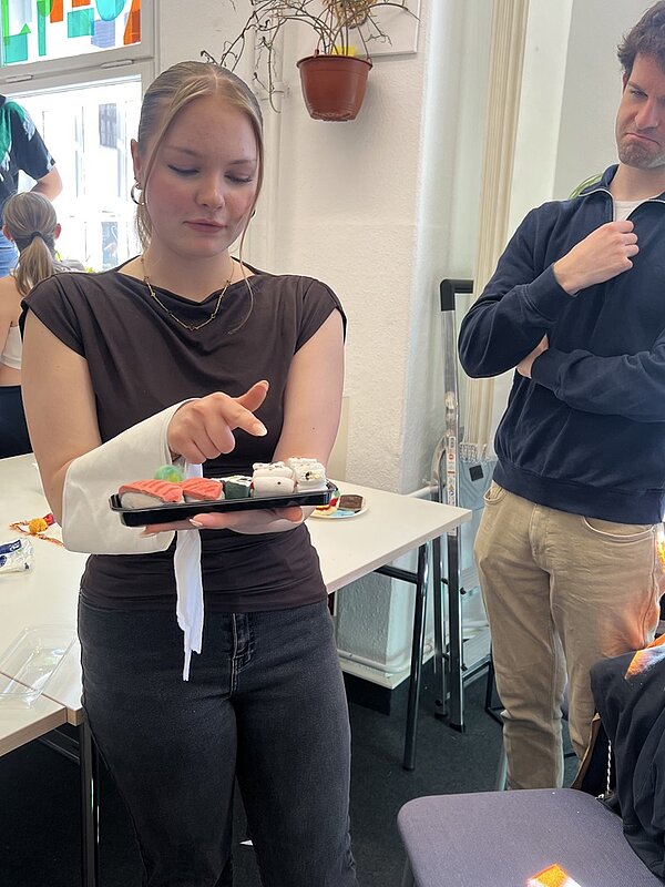 Eine Auszubildende mit verbundenem rechten Arm hält ein Tablett mit Sushi in der linken Hand und zeigt mit dem Zeigefinger der rechten Hand auf das Sushi. Sie trägt ein dunkles T-Shirt und eine helle Hose. Im Hintergrund steht ein Azubi mit verschränkten Armen und nachdenklichem Gesichtsausdruck. Im Raum befinden sich weiße Tische, Stühle und eine an der Wand hängende Pflanze in einem Blumentopf. Durch ein Fenster fällt Tageslicht in den Raum.