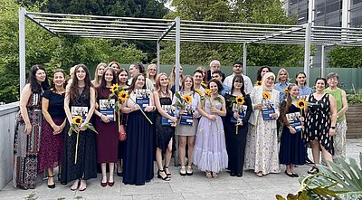 Eine Gruppe von festlich gekleideten Menschen steht im Freien unter einem Pavillon und hält Zertifikate und Sonnenblumen. Sie lächeln und posieren für ein Gruppenfoto. Im Hintergrund sind Bäume und ein Gebäude zu sehen.
