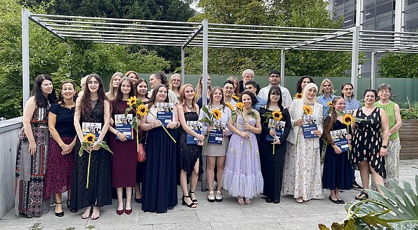 Eine Gruppe von festlich gekleideten Menschen steht im Freien unter einem Pavillon und hält Zertifikate und Sonnenblumen. Sie lächeln und posieren für ein Gruppenfoto. Im Hintergrund sind Bäume und ein Gebäude zu sehen.