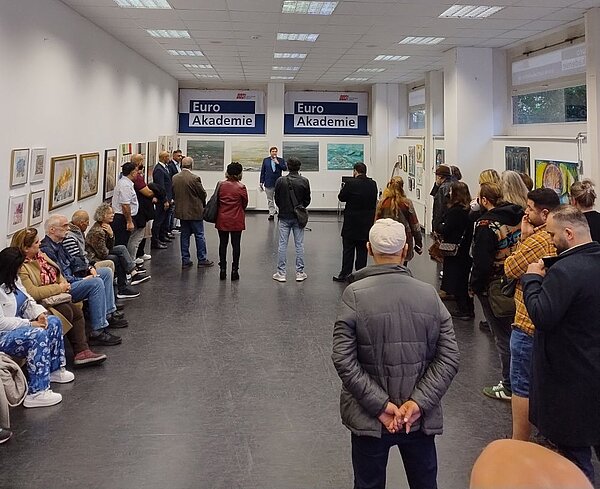 Heller Ausstellungsraum mit weißen Wänden und zahlreichen gerahmten Kunstwerken. Besucherinnen und Besucher hören einem Mann zu, der in eine Mikrophon spricht. Im Hintergrund hängen Banner mit der Aufschrift „Euro Akademie“.