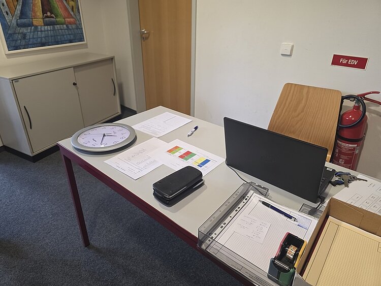 Abschlussprüfung im Fach Mathematik an der Euro Akademie Dresden Auf dem Lehrerpult der Lehrkraft steht ein aufgeklappter Laptop, eine große runde Wanduhr, Kugelschreiber und Arbeitsblätter zur Prüfung sowie ein Schlüsselbund und eine Aktenablage. Im Hintergrund sieht man einen Feuerlöscher, eine Tür aus Holz, ein buntes, großformatiges Bild hängt an der Wand, darunter steht ein kleiner heller Schrank.