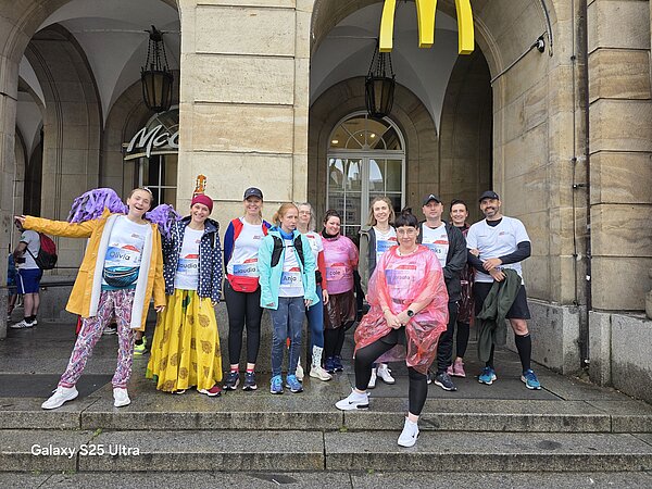 Gruppenfoto vor einem McDonald's-Bogen mit zwölf Teilnehmenden der REWE Team Challenge, darunter viele mit Startnummern und Namensschildern.