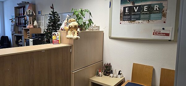 Ein weihnachtlich geschmückter Empfangsbereich mit einer hellen Theke und einem kleinen Tisch mit zwei Holzstühlen. Auf der Theke steht eine kleine Tanne mit Weihnachtskugeln, daneben ein Plüschtier in Form eines Teddybären mit rotem Schal und Weihnachtsmütze. Im Hintergrund ist ein größerer Weihnachtsbaum mit Lichterketten und Kugeln zu sehen. An der Wand hängt ein gerahmtes Poster mit dem Schriftzug 'EVER' und einem kleinen Schild mit der Aufschrift 'Euro Schulen'. Auf dem kleinen Tisch stehen weitere weihnachtliche Dekorationen, darunter eine kleine Figur und ein Miniatur-Weihnachtsbaum. Die Räumlichkeiten wirken freundlich und festlich geschmückt, passend zur Weihnachtszeit in den Euro-Schulen und Euro Akademien in Dresden, Meißen und Riesa.