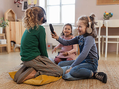 Drei Kinder sitzen auf dem Boden in einem hellen Raum. Zwei der Kinder tragen Kopfhörer und eines hält ein Mikrofon in der Hand. Sie scheinen gemeinsam Musik zu hören oder zu singen und wirken fröhlich und engagiert.