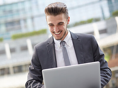 Ein junger Mann im Business Look - Anzug mit grauem Jacket, weißem Hemd und silberfarbener Krawatte - sitzt freudig lächelnd vor einem weißem  Laptop.