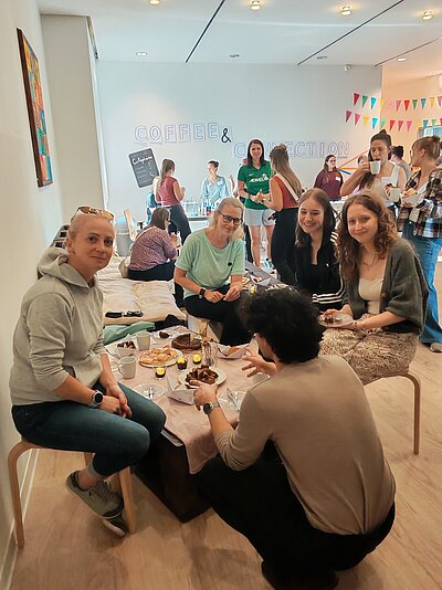 Mehrere Personen sitzen und stehen bei einem gemeinsamen Treffen mit Kaffee und Gebäck in einem hellen Raum. An der Wand steht die Aufschrift „Coffee & Connection“.